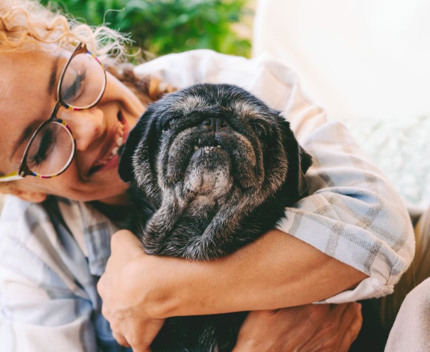 A woman hugging a black pug, both appearing joyful and content.